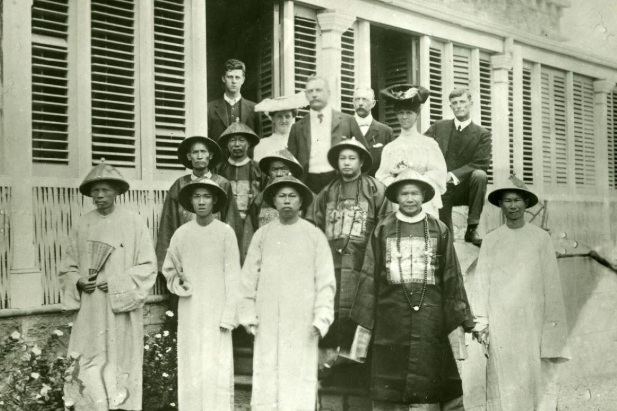 Black and white photo, several people standing in front of a house. Many Chinese merchants dressed in traditional attire. 
