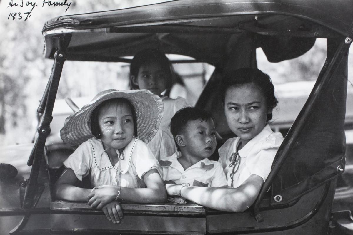 Black and white photo. Two children and a woman sitting in the front seat of an older style car. Women on the seat and two children on leaning on the dashboard. All looking out the passenger side door. 