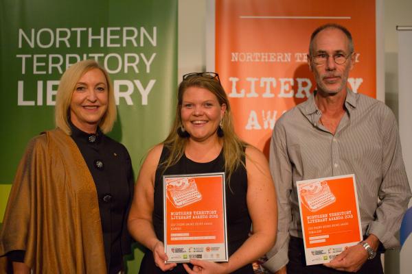 Two women and a man standing side by side. The woman and man on the right are holding orange awards certificates