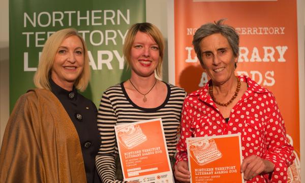 Three women standing side by side. The two women on the right are holding orange awards certificates