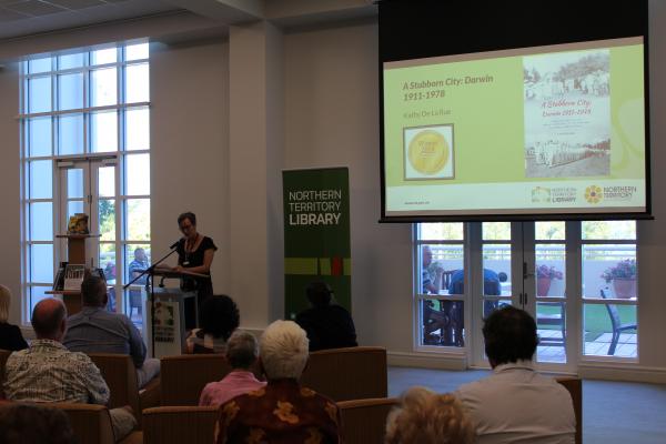 Woman at lectern reading a speech. In the background a green pull up banner and projection screen with the winning work displayed. 