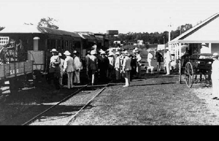 Black and white photograph of men standing and waiting at a train station. Men wearing white safari style hats and white suits.