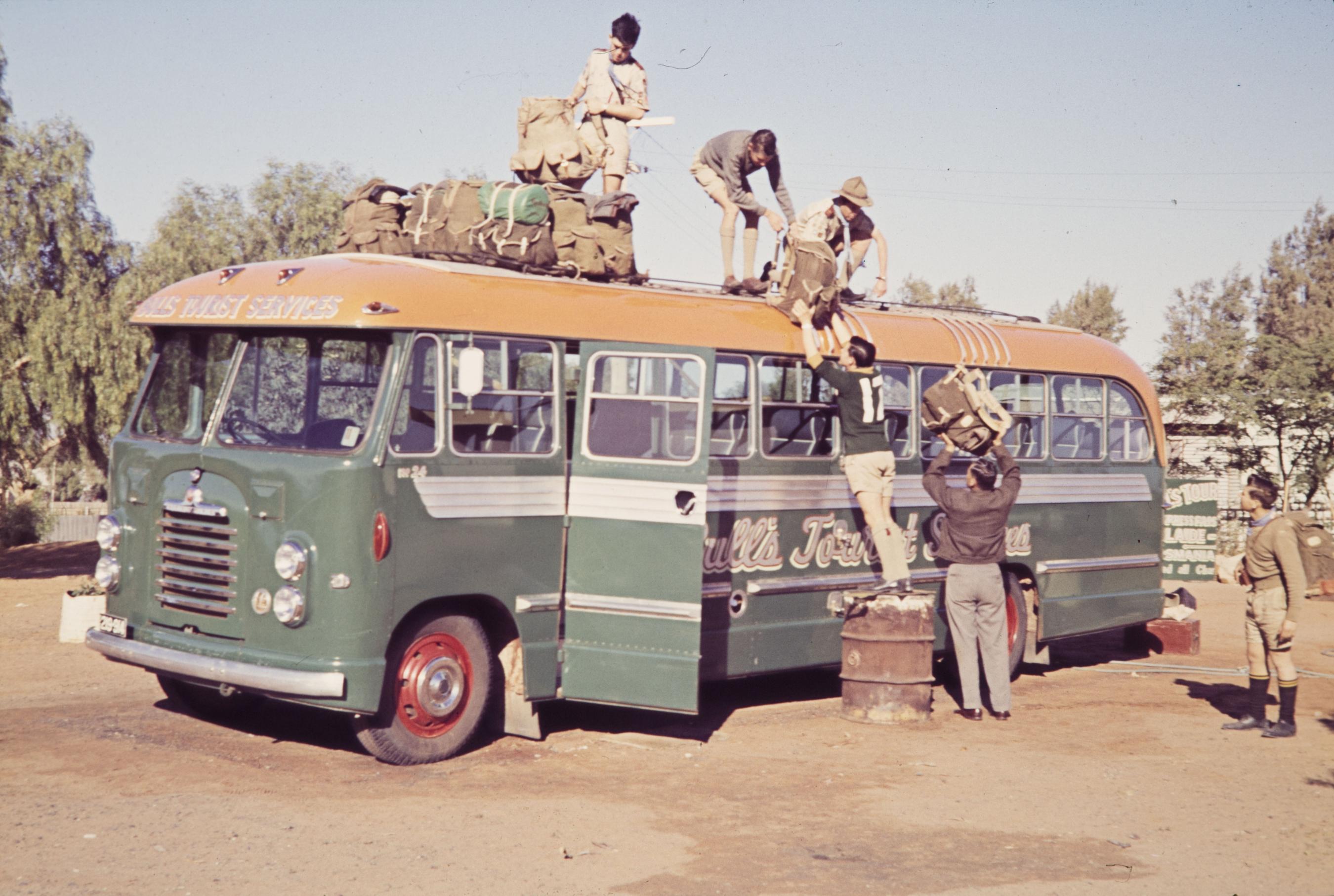 People packing a green and orange tour bus circa 1965. People standing on the roof of the bus and others passing luggage up from the ground. 