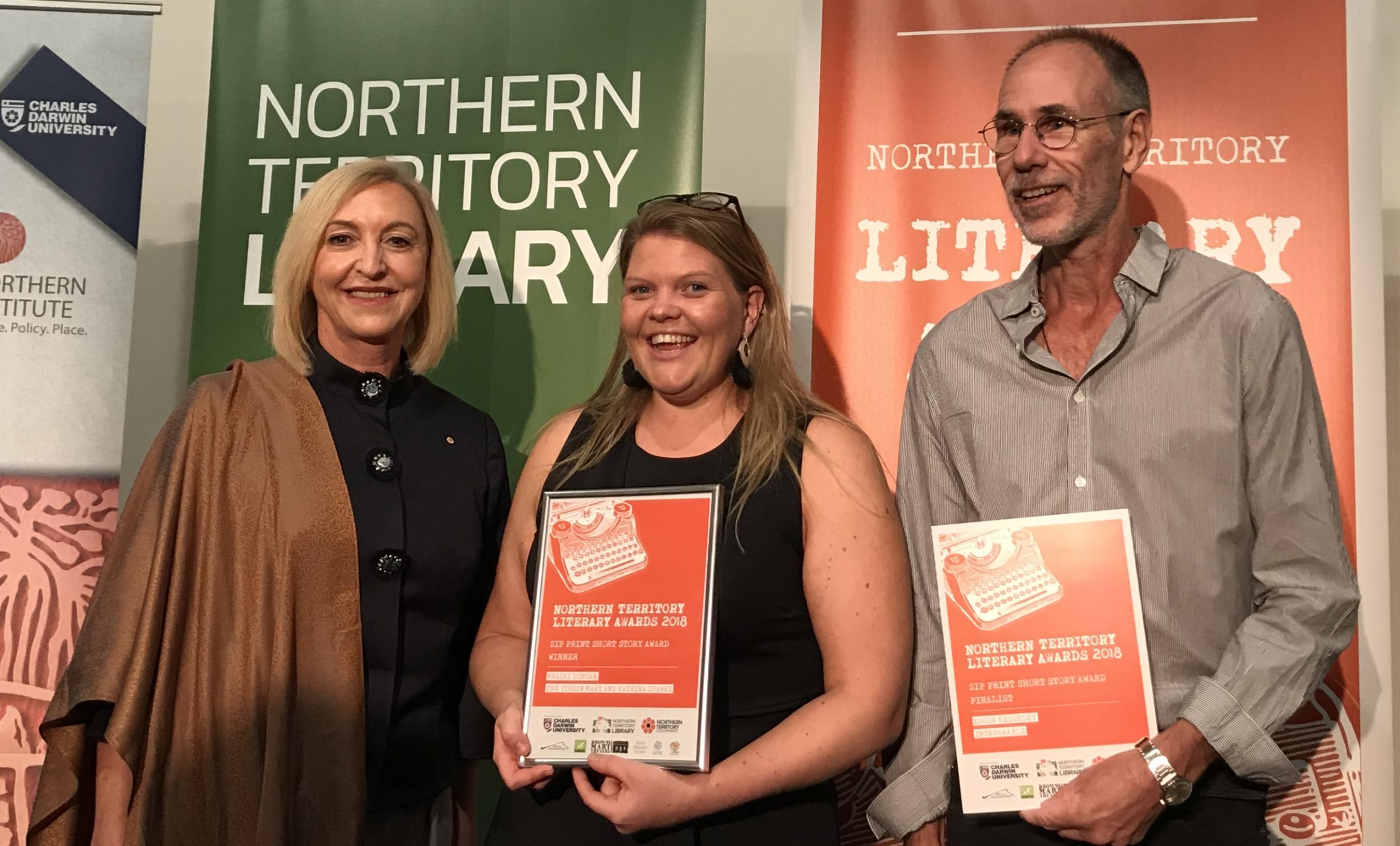 Her Honour the Hounourable Vicki O'Halloran Administrator of the Northern Territory with Mharii Duncan winner of the Short Story Award and Edgar Keighley finalist in the Poetry Award. Event banners in the background. 