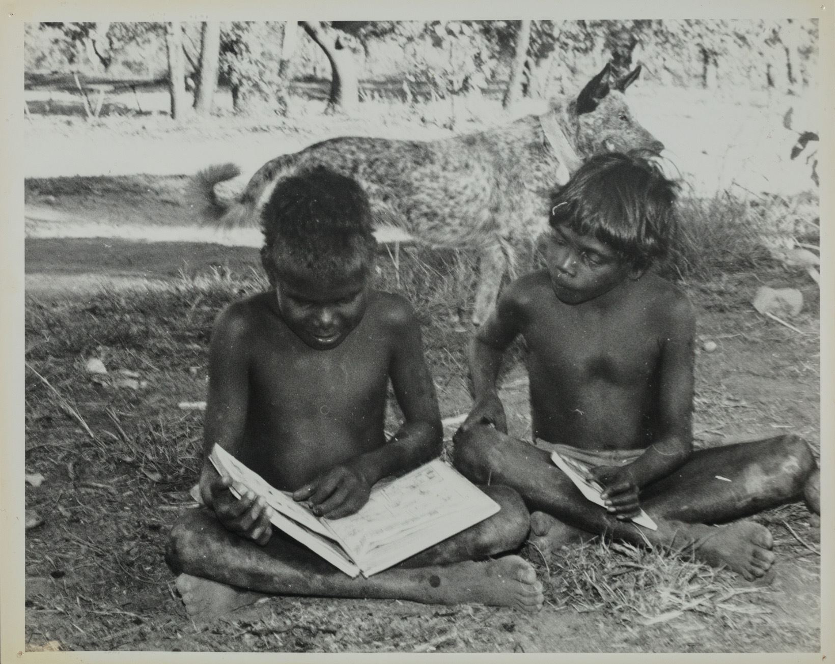 Two young boys with no shirts on sitting crossed legged on the ground reading books