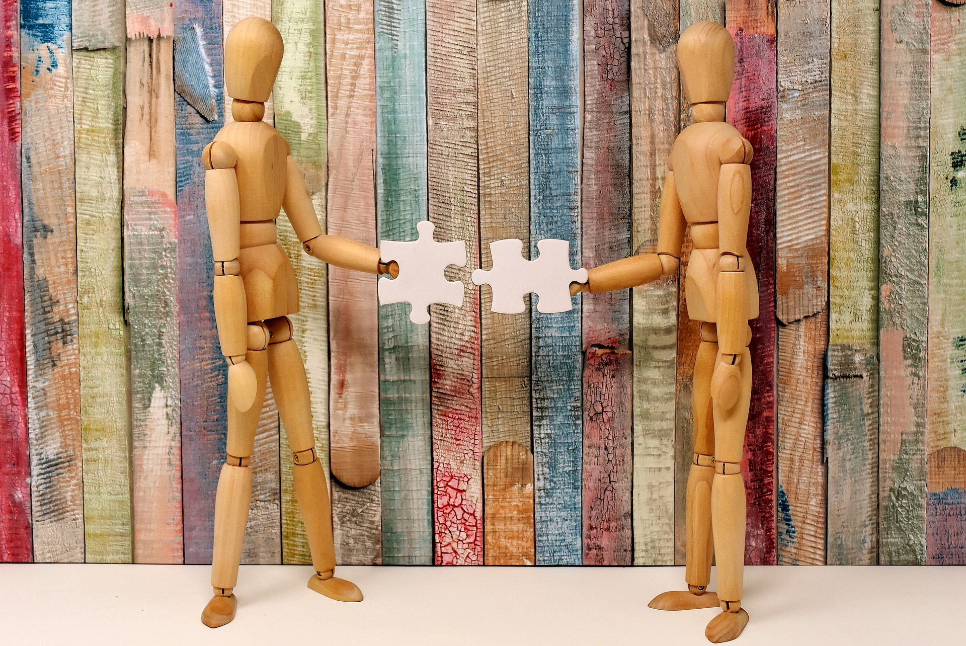 Two wooden drawing models standing in front of coloured panel fencing made of paddle pop sticks each holdig a piece of a puzzle and coming together