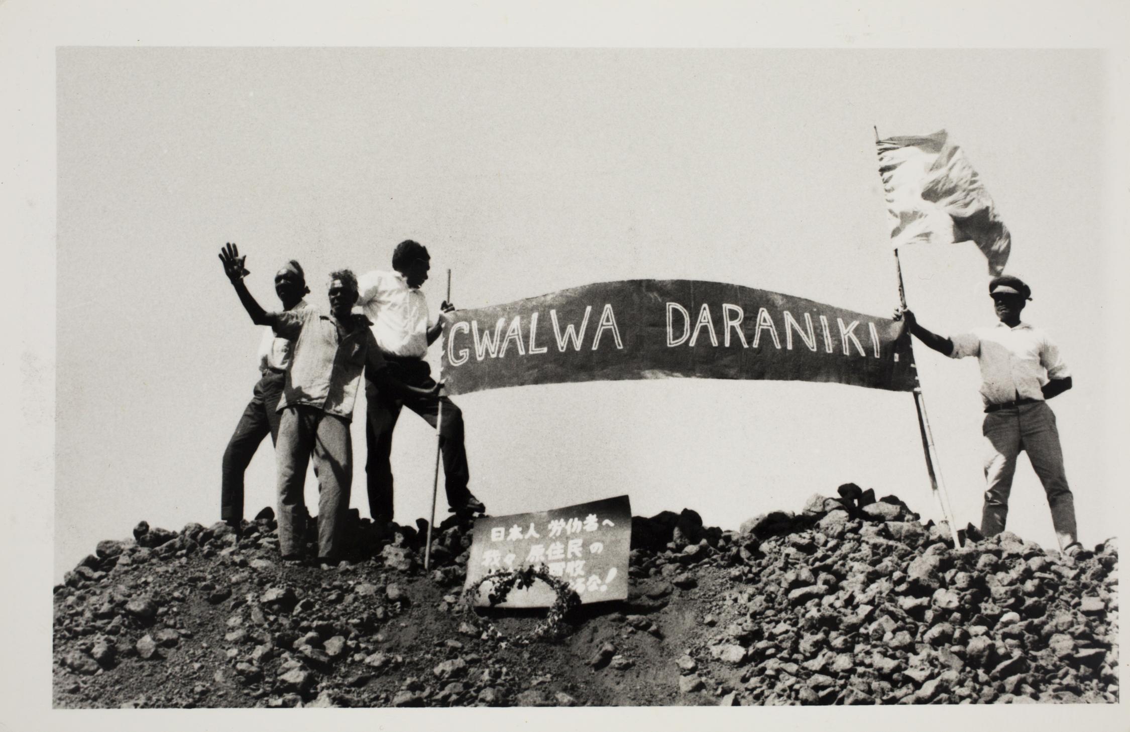 Four men standing on top a mountain holding a protest sign that reads "Gwala Daraniki" meaning "Our Land"