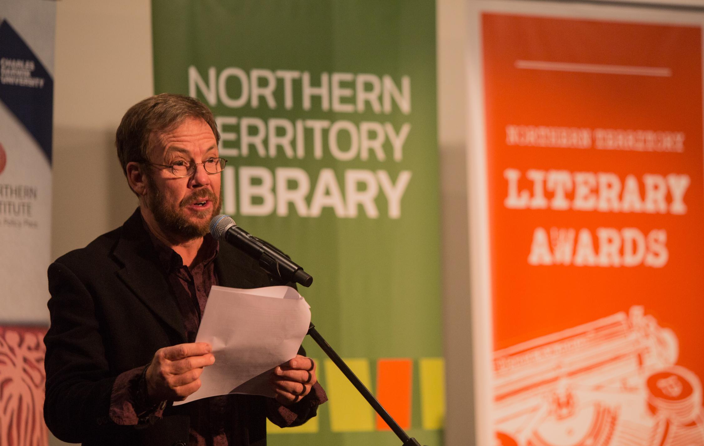 Man on left hand side wearing glass, dark jacket and shirt reading into a microphone. Behind are green and orange banners. 