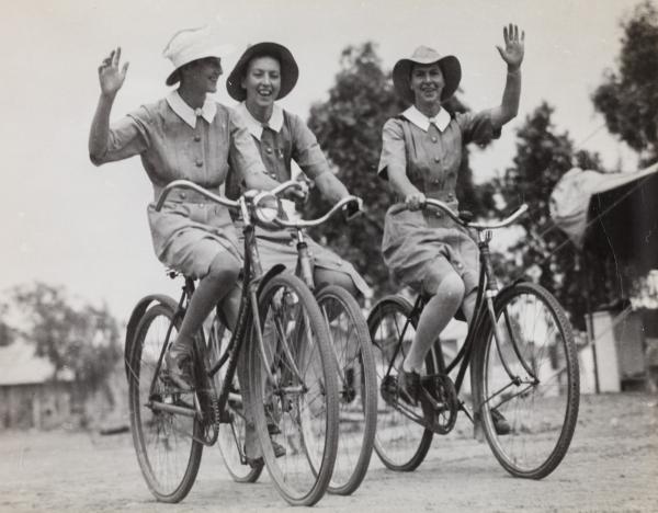 Three nurses in uniform riding bicycles and waving to the camera.
