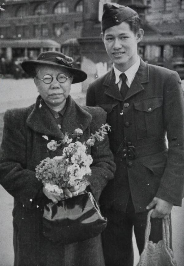 Black and white photo. Young man in RAAF uniform stands next to elderly woman holding a bouquet of flowers.