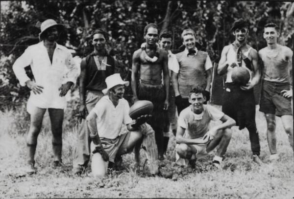 Nine men of various ethnicities, casually standing and kneeling, one holding a football and another a basketball. All wearing one or two pieces of formal wear or other fancy dress. 